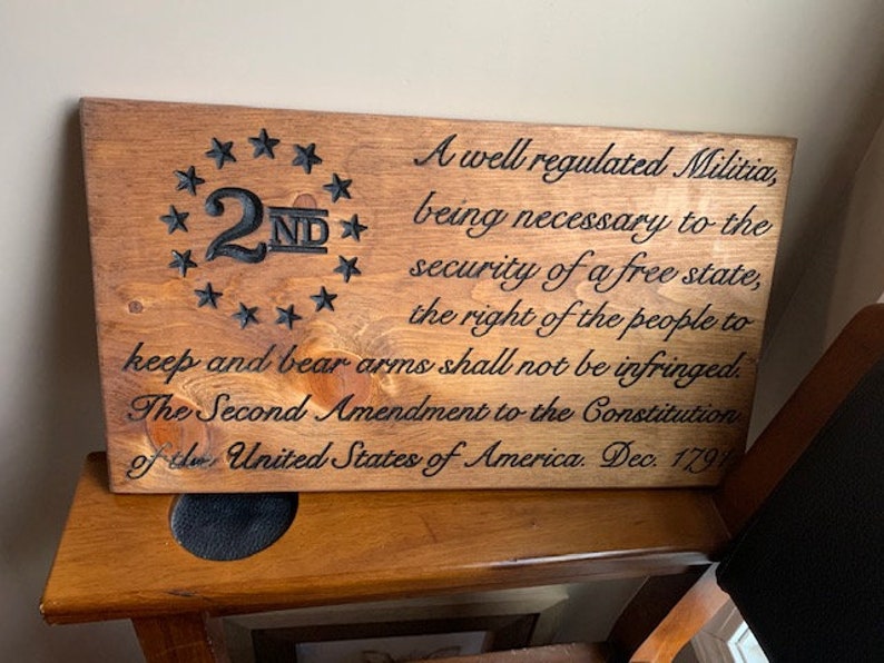 Puede incluir: Un letrero de madera con el texto "A well regulated Militia, being necessary to the security of a free state, the right of the people to keep and bear arms shall not be infringed. The Second Amendment to the Constitution of the United States of America. Dec. 1791" y un c&iacute;rculo con estrellas alrededor del n&uacute;mero "2ND" en el centro.