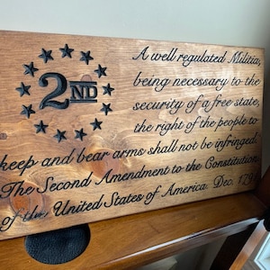 Könnte beinhalten: Ein Holzschild mit dem Text "A well regulated Militia, being necessary to the security of a free state, the right of the people to keep and bear arms shall not be infringed. The Second Amendment to the Constitution of the United States of America, Dec. 1791" und einem sternförmigen Design mit der Nummer "2ND" in der Mitte.