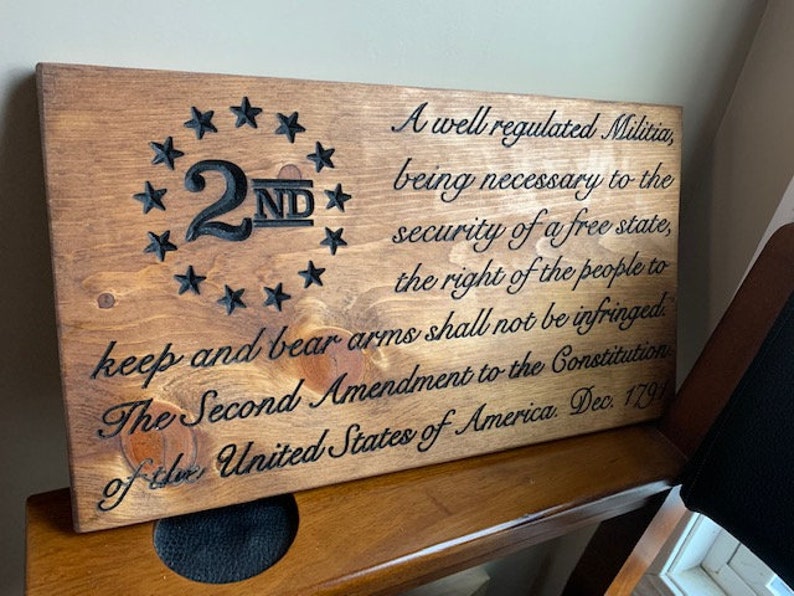Puede incluir: Un letrero de madera con el texto "A well regulated Militia, being necessary to the security of a free state, the right of the people to keep and bear arms shall not be infringed. The Second Amendment to the Constitution of the United States of America, Dec. 1791" y un dise&ntilde;o en forma de estrella con el n&uacute;mero "2ND" en el centro.