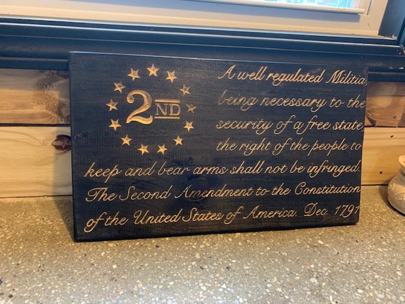 Puede incluir: Un letrero de madera negro con letras doradas. El letrero presenta el texto "2ND" con un c&iacute;rculo de estrellas alrededor. El texto "A well regulated Militia, being necessary to the security of a free state the right of the people to keep and bear arms shall not be infringed. The Second Amendment to the Constitution of the United States of America. Dec. 1791" tambi&eacute;n est&aacute; en el letrero.