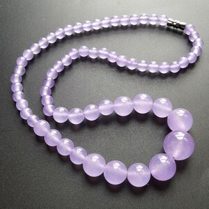 May include: A necklace featuring round, lavender-colored beads. The beads graduate in size, with larger beads at the center and smaller ones towards the clasp. The clasp is silver-colored. The necklace is displayed against a dark background.