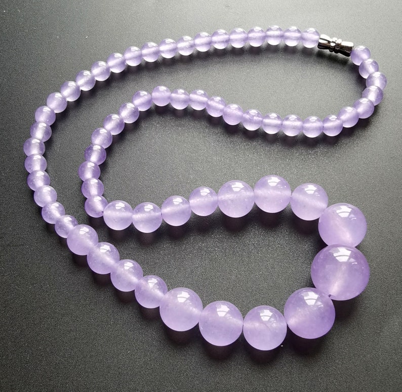 May include: A necklace featuring round, lavender-colored beads. The beads graduate in size, with larger beads at the center and smaller ones towards the clasp. The clasp is silver-colored. The necklace is displayed against a dark background.