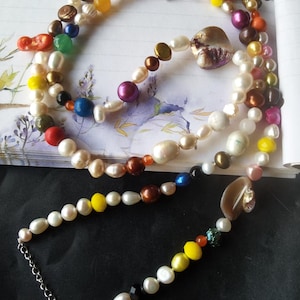 Puede incluir: Un collar de múltiples hilos con una variedad de perlas y cuentas de colores. El collar incluye cuentas blancas, amarillas, naranjas, rojas, verdes, azules y moradas, con un cierre de cadena plateada. Las perlas varían en tamaño y forma.