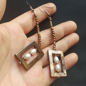 May include: A pair of long, brown beaded earrings with a rectangular, brown, and white patterned frame. The frame holds three white pearls.