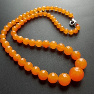 May include: A necklace featuring polished, round, orange beads. The beads graduate in size, with larger beads at the center and smaller ones towards the clasp. The clasp is silver-colored. The necklace is displayed on a dark surface.