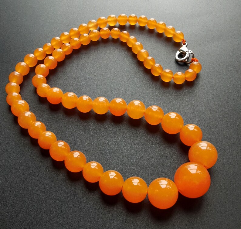 May include: A necklace featuring polished, round, orange beads. The beads graduate in size, with larger beads at the center and smaller ones towards the clasp. The clasp is silver-colored. The necklace is displayed on a dark surface.