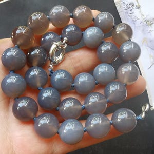 May include: A necklace featuring large, round, gray beads strung on blue cord. The beads have a polished, reflective surface. A silver-colored clasp secures the necklace. The necklace is displayed against a neutral background.