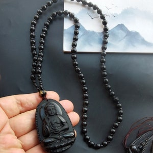May include: Black obsidian Buddha pendant necklace with a black beaded chain. The pendant features a detailed carving of a seated Buddha.