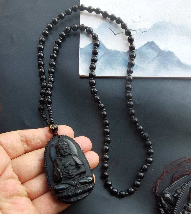 May include: Black obsidian Buddha pendant necklace with a black beaded chain. The pendant features a detailed carving of a seated Buddha.