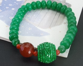 May include: A green jade bracelet with a carved green jade bead and a red agate bead. The bracelet is made of round beads and is strung on a clear elastic cord.