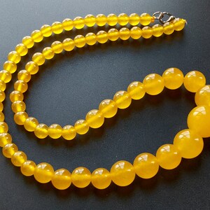 May include: A necklace featuring translucent, round, yellow beads. The beads graduate in size, with larger beads at the front and smaller ones towards the clasp. The necklace is displayed against a dark background, highlighting the beads' color and shine.