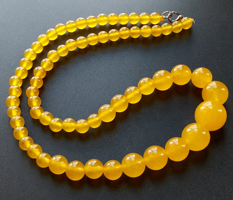 May include: A necklace featuring translucent, round, yellow beads. The beads graduate in size, with larger beads at the front and smaller ones towards the clasp. The necklace is displayed against a dark background, highlighting the beads' color and shine.
