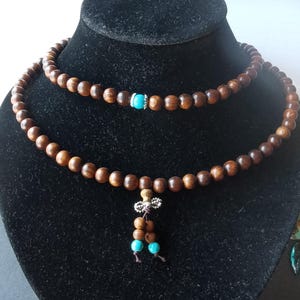 May include: A brown wooden mala necklace with turquoise beads and a silver charm. The necklace is displayed on a black velvet stand.