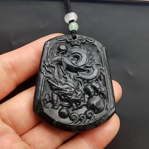 May include: A black jade pendant with a detailed carving of a dragon. The pendant is attached to a black cord with green and white beads.
