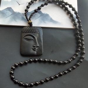 May include: Black beaded necklace with a black carved Buddha pendant. The pendant is rectangular and features a detailed carving of a Buddha's face.
