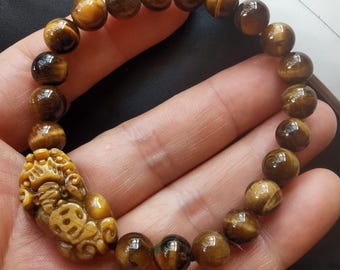 Op de afbeelding: Een bruine tijgeroog-parel armband met een gegraveerde tijgeroog-bedel. De bedel heeft de vorm van een mythisch wezen met ingewikkelde details.