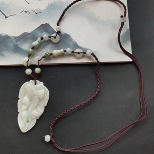 Puede incluir: Un colgante de jade blanco tallado en forma de hoja con un collar de cordón trenzado marrón con cuentas verdes y blancas.