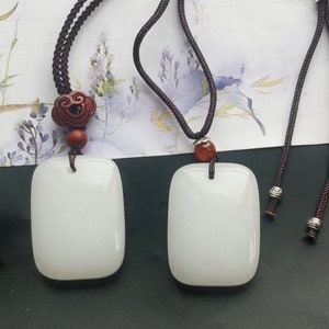 Könnte beinhalten: Zwei weiße Jade-Anhänger an braunen Schnüren mit dekorativen Perlen. Der Anhänger links hat eine Holzperle mit einem geschnitzten Design. Der Anhänger rechts hat eine rote Achatperle.