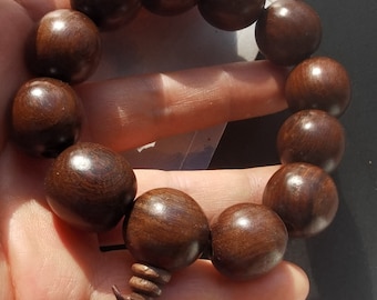 Op de afbeelding: Een donkerbruine houten kralen armband met een geknoopte kwast. De kralen zijn rond en glad, en de armband is geregen op een donkerbruin koord.