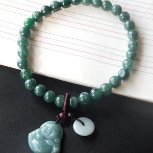 Op de afbeelding: Een groene jade armband met een jade Boeddha-bedel en een jade muntbedel. De armband is gemaakt van ronde kralen en heeft een donkerbruin koord.