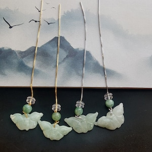 Pode incluir: Quatro pares de brincos delicados. Cada brinco apresenta um pingente de jade em forma de borboleta, uma conta verde e uma conta transparente. Dois pares têm correntes douradas e dois têm correntes prateadas. O fundo é uma paisagem montanhosa em aquarela.