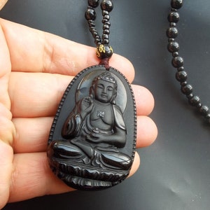 May include: Black obsidian pendant necklace featuring a detailed carving of a seated Buddha. The necklace is made of black beads and has a gold-colored accent.