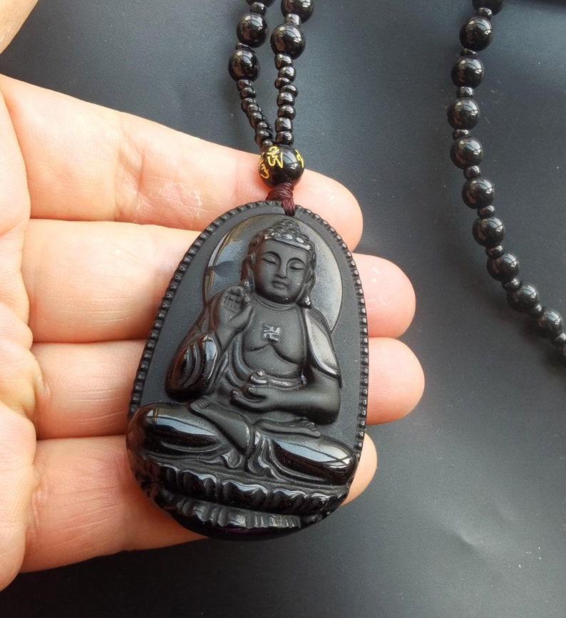 May include: Black obsidian pendant necklace featuring a detailed carving of a seated Buddha. The necklace is made of black beads and has a gold-colored accent.