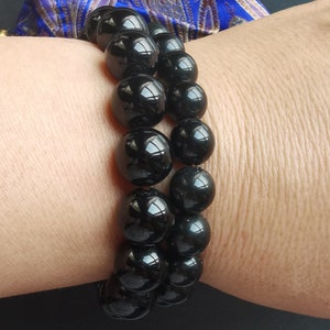May include: Two black beaded bracelets. The bracelets are made of large, glossy, round beads. The beads reflect light, creating highlights. The bracelets are worn on a wrist, with a glimpse of a blue and gold patterned fabric in the background.