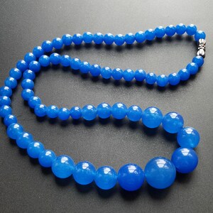 May include: A necklace of graduated, round, bright blue beads. The beads vary in size, with the largest at the center and smaller ones towards the ends. A silver-colored clasp secures the necklace. The beads have a glossy finish.