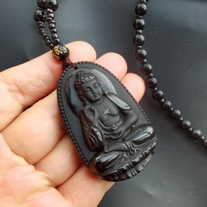 May include: Black obsidian pendant necklace featuring a carved Buddha figure. The necklace is made of black beads and has a brown cord.
