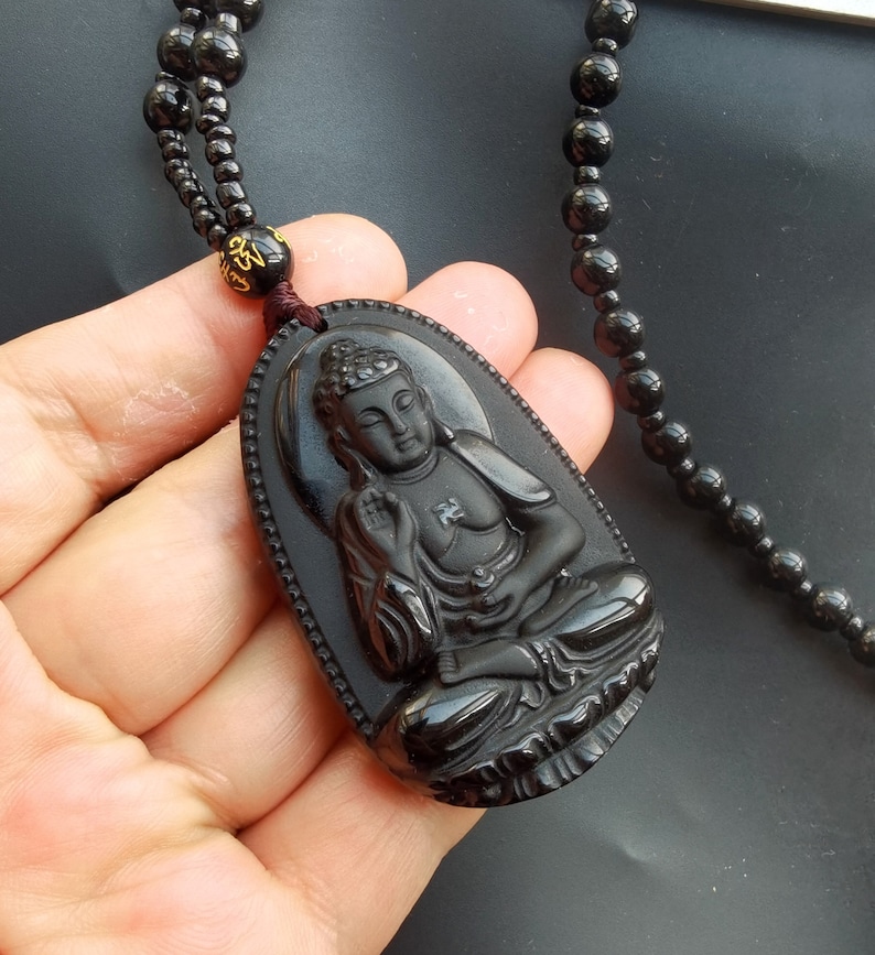 May include: Black obsidian pendant necklace featuring a carved Buddha figure. The necklace is made of black beads and has a brown cord.
