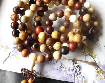 Puede incluir: Un collar de cuentas de madera en varios colores, incluyendo crema, marrón, rojo y negro. Las cuentas redondas están ensartadas, con un nudo decorativo y cuentas adicionales al final. Las cuentas son probablemente para meditación o joyería.