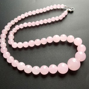 May include: A necklace composed of polished, light pink rose quartz beads. The beads vary in size, with larger spheres at the front and smaller ones towards the clasp. The necklace is strung on a visible cord and secured with a silver-colored clasp.