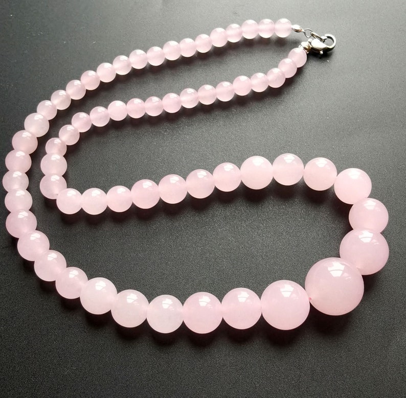 May include: A necklace composed of polished, light pink rose quartz beads. The beads vary in size, with larger spheres at the front and smaller ones towards the clasp. The necklace is strung on a visible cord and secured with a silver-colored clasp.
