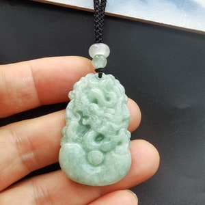 May include: A green jade pendant carved with a detailed dragon design. The pendant is attached to a black cord with a white bead.