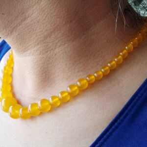 May include: A golden yellow beaded necklace with graduated sizes. The necklace is made of round beads and is worn around the neck. The background is a blue top and skin.