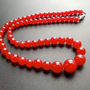 May include: A vibrant red beaded necklace. The necklace features large, round beads that graduate in size, strung together with a silver clasp. The beads have a glossy finish, reflecting light. The necklace is displayed against a dark background.