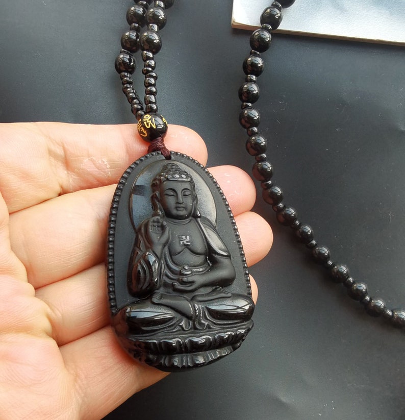 May include: Black obsidian Buddha pendant necklace with a black beaded chain. The pendant features a detailed carving of a seated Buddha with a gold inscription on the chest.