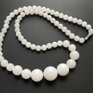 May include: A white beaded necklace with varying sized round beads. The necklace has a magnetic clasp. The beads are a bright white color and appear to be smooth. The necklace is displayed on a black surface.