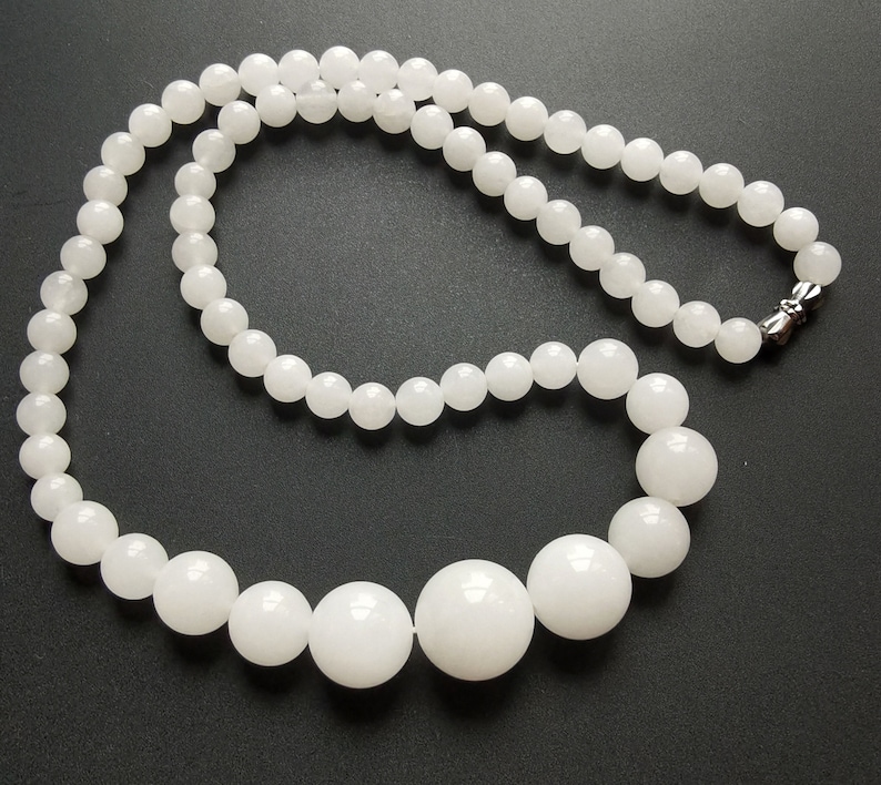 May include: A white beaded necklace with varying sized round beads. The necklace has a magnetic clasp. The beads are a bright white color and appear to be smooth. The necklace is displayed on a black surface.
