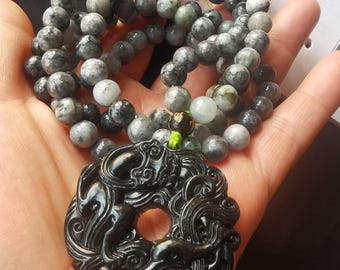 May include: A beaded necklace with a black carved pendant. The necklace features gray and white round beads. The pendant is a dark, intricately carved design with a central hole. The necklace is held in a hand.