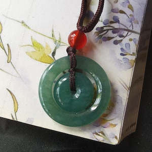 Puede incluir: Un collar con un colgante de jade verde con un diseño de doble círculo. El colgante está unido a un cordón marrón con una cuenta roja.