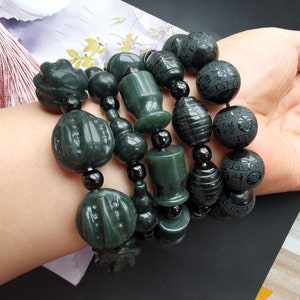 Pode incluir: Um bracelete de jade com contas pretas e pingentes de jade talhados. Os pingentes incluem uma cabaça, um pêssego e uma flor. O bracelete está no pulso de uma pessoa.