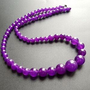 May include: A necklace featuring graduated, round beads in a deep purple hue. The beads vary in size, with the largest ones at the front and smaller ones towards the clasp. The necklace is displayed against a dark background.