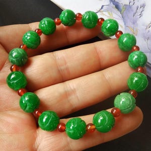 May include: A jade bracelet featuring carved green beads interspersed with small, round, reddish-orange beads. The jade beads have a floral design. The bracelet is displayed on a hand.