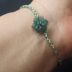 Op de afbeelding: Een armband met een groene jade bloemenbedel aan een gevlochten koord van goud en groen.