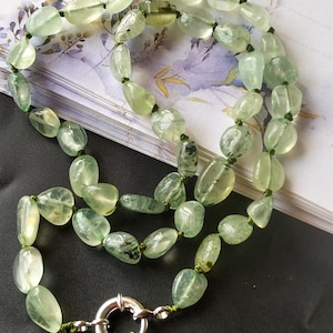 May include: A necklace featuring light green, oval-shaped beads strung together with green thread. The beads have a translucent quality and are secured with a silver-colored clasp. The necklace is displayed on a patterned surface.
