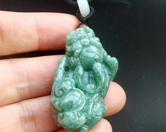Puede incluir: Un colgante de jade verde tallado en forma de criatura mítica con detalles intrincados. El colgante está unido a un cordón negro.