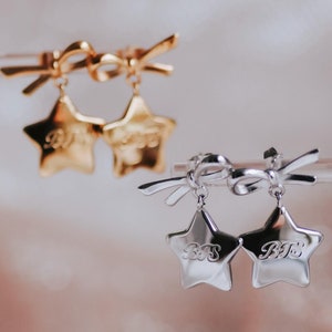 May include: A pair of star-shaped earrings with bows. The earrings are made of gold and silver metal. The stars have the letters "BTS" engraved on them.