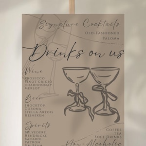 May include: A tan-colored sign on an easel with the words "Drinks on us" in elegant script. The sign lists drink options: wine, beer, spirits, and non-alcoholic beverages. Decorative drawings of glasses and text are included.